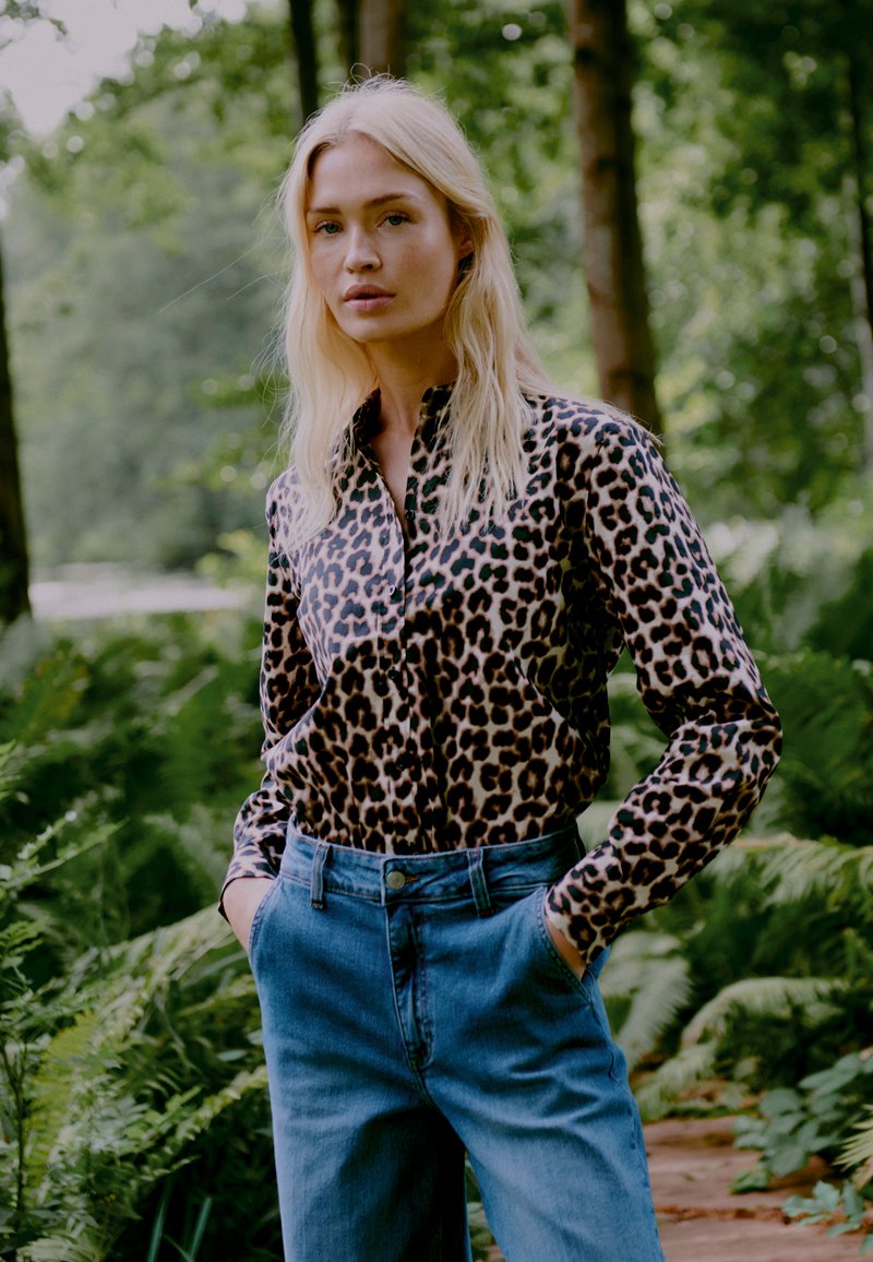 Košile s leopardím potiskem na knoflíky vyrobená z hedvábné tkaniny, spárovaná s vysokými modrými džíny. Model stojící s rukama v kapsách, v pozadí listnatá krajina.