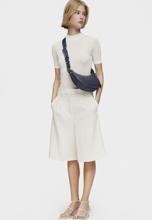 Borsa a tracolla blu con logo in rilievo, realizzata in materiale liscio. Il modello indossa una maglietta bianca aderente e pantaloni corti plissettati color crema con sandali flat.