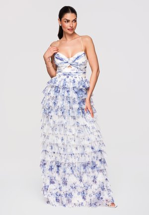 Femme portant une robe maxi à volants blanche et bleue à fleurs, avec des bretelles fines, posant avec une main touchant la robe et l’autre près du cou.