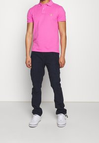 Rosa polo T-shirt med två knappar och en liten logotyp, tillsammans med mörka cargobyxor och vita sneakers. Outfiten har en avslappnad stil.