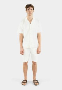 Weißes Kurzarmhemd mit Kragen und passende Shorts aus strukturiertem Stoff. Das Outfit wird mit braunen Sandalen kombiniert.