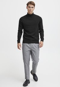 Maglione a collo alto nero con maniche lunghe, abbinato a pantaloni grigi chiari su misura. Il modello indossa scarpe nere, posando su uno sfondo neutro.