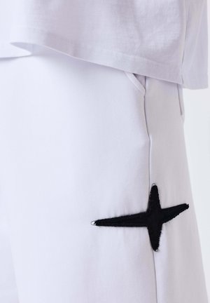 Close-up van witte broek met zijzak en zwarte geborduurde ster bij de zak, gecombineerd met een witte zoom van het overhemd.