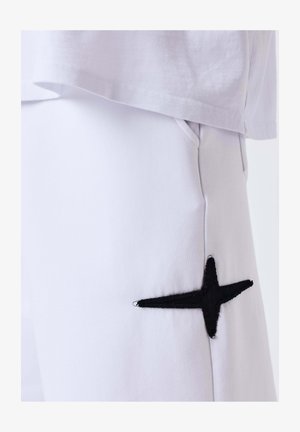 Primo piano di pantaloni bianchi con tasca laterale e stella nera ricamata vicino alla tasca, abbinati a un orlo di camicia bianca.