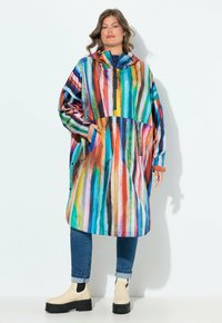 Imperméable surdimensionné coloré avec des rayures verticales bleues, roses et vertes. Comprend une capuche, une poche avant et un détail de fermeture éclair. Porté par-dessus un jean.