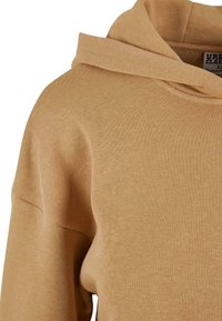 Sweat à capuche en coton beige avec capuche visible et couture d'épaule, montrant la texture et une petite partie d'une étiquette de vêtement à l'intérieur du col.