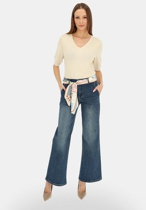 Kobieta stojąca z rękami w kieszeniach, ubrana w beżową bluzkę z krótkim rękawem, szerokie niebieskie dżinsy, wzorzysty szalik jako pasek oraz beżowe szpilki na białym tle.