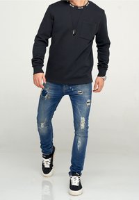 Hombre vestido con sudadera negra, vaqueros azules desgastados, zapatillas negras con cordones blancos y un collar de perro negro, caminando sobre un suelo reflectante.