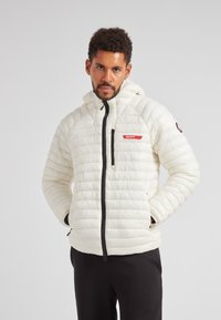 Giacca a vento bianca con motivo trapuntato orizzontale, chiusura con zip e cappuccio regolabile. Presenta un marchio rosso e cerniere nere.