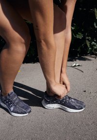 Atletische schoenen met een zwart en grijs gebreid bovenwerk, een wit gemêleerde zool, flexibele ontwerp en vetersysteem, worden vastgemaakt op een voetgangersoppervlak.