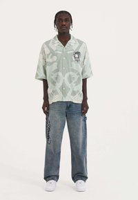 Green patterned short-sleeve shirt with a lion graphic; paired with light blue baggy jeans featuring black lettering; white sneakers.