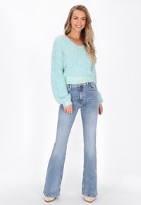 Hellblauer, flusiger Strickpullover mit V-Ausschnitt und geripptem Saum, kombiniert mit hellen Schlagjeans und beigen Plateauschuhen.