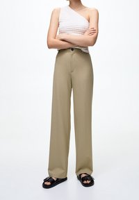 Top crema texturizzato a spalla unica abbinato a pantaloni khaki a vita alta e gamba larga, con sandali neri flat. Tessuto liscio, dettagli minimi.