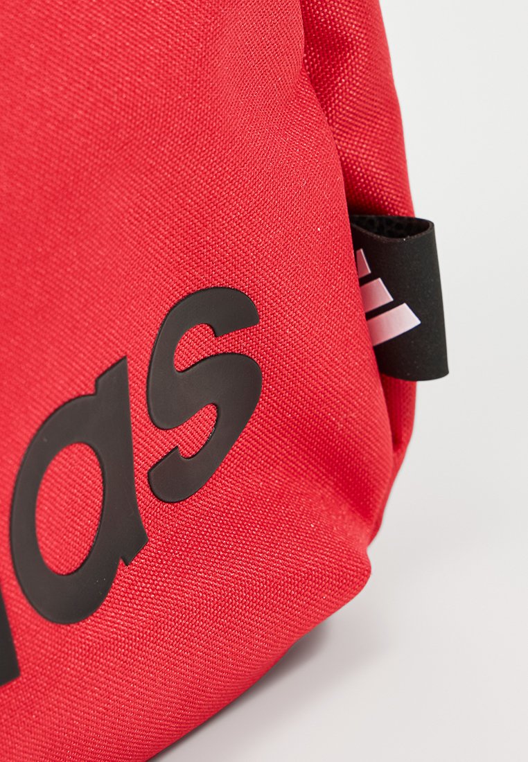 Tela roja con texto parcial en relieve en negro que dice "das" y una pequeña etiqueta negra con el logo blanco de Adidas en el lateral.