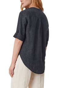 Femme portant une chemise en lin gris foncé aux manches retroussées et un pantalon beige, vue de dos sur fond blanc.