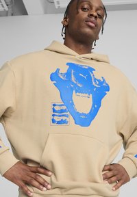 Felpa beige con una grande grafica blu di un viso simile a un teschio e il testo che recita “PUMA HOOPS.” Tessuto morbido, tasca frontale e polsi a coste.