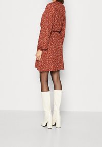 Vestido floral rojo con mangas largas, cintura ceñida y falda fluida. Combinado con botas de tacón alto brillantes y hasta la rodilla en color blanco.