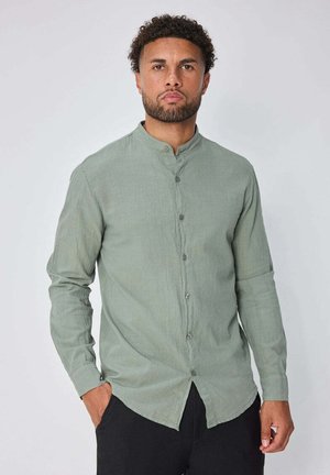 Homme aux cheveux courts et bouclés avec une barbe, porte une chemise vert clair à manches longues avec un col mandarin et un pantalon noir, debout avec une main dans la poche.