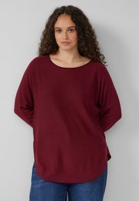 Bordeauxfarbener Strickpullover mit weitem, rundem Ausschnitt und langen Ärmeln; hat eine lässige Passform und einen abgerundeten Saum, kombiniert mit blauen Jeans.