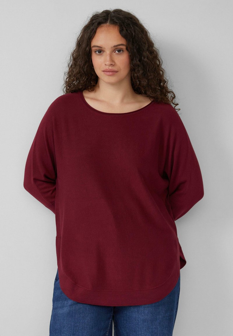 Bordeauxfarbener Strickpullover mit weitem, rundem Ausschnitt und langen Ärmeln; hat eine lässige Passform und einen abgerundeten Saum, kombiniert mit blauen Jeans.