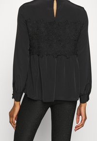 Czarna bluzka z koronkowym panelem, długimi rękawami z zapięciem na guziki oraz otwarciem w kształcie łezki z tyłu. Miękki, lekko opadający materiał.