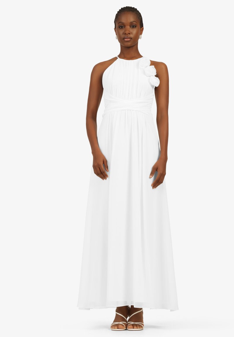 Weißes Maxikleid mit Neckholder und gerafftem Oberteil, verziert mit Stoffblumen an einer Schulter und einem fließenden Rock. Getragen mit Riemchensandalen.