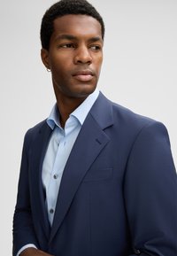 Mann in navyblazer und hellblauem Hemd, der mit neutralem Ausdruck zur Seite schaut, vor einem schlichten Hintergrund.