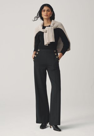 Černé široké kalhoty s detaily zlatých knoflíků a bočními kapsami, doplněné černým svetrem a světle béžovým pleteným šálem.