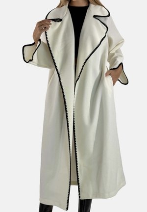 Lange witte jas met zwarte biezen, voorzien van een brede kraag, open ontwerp en grote zijzakken, gemaakt van een gestructureerde stof.