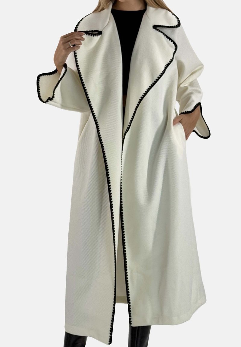 Lang hvid frakke med sort kant, med en bred krave, åben design og store sidelommer, lavet af tekstureret stof.