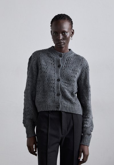 Cardigan gris tricoté avec un col rond, fermeture à boutons et motifs texturés. Manches côtelées et ourlet, associé à un pantalon noir.