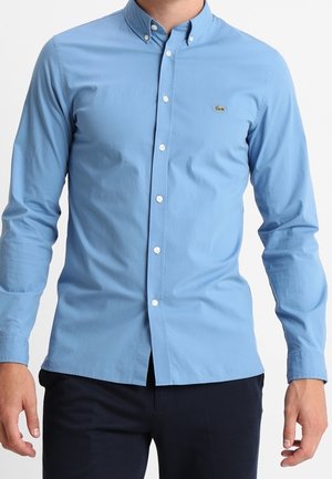 Man draagt een lichtblauw overhemd met lange mouwen en knopen, met een klein krokodillogo op de borst, gecombineerd met een donkere broek.
