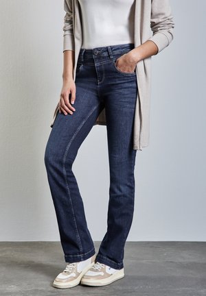 Jeans bleu foncé avec une jambe droite, un design à cinq poches, et une texture en denim lisse, associés à des baskets claires et un cardigan beige.