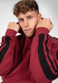 Bordeaux sweatshirt met zwarte accentstrepen, capuchon en trekkoord. Bevat geborduurd logo op de borst en heeft een zachte textuur.