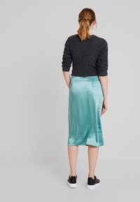 En bakvy av en person som bär en svart stickad tröja och en ljus teal satin midi kjol, tillsammans med svarta och vita sneakers.