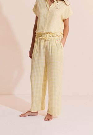 Vrouw draagt een top met korte mouwen in geel-wit geruit patroon en losse broek met kleine kersenborduursels, staat op blote voeten met één hand in de zak.