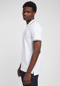 Uomo con capelli ricci corti che indossa una polo bianca con bordi neri e jeans scuri, in piedi di profilo su uno sfondo semplice.