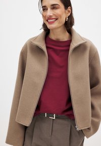 Bežová zipsová bunda s vysokým golierom, vyrobená z jemnej látky, oblečená cez vinsovú tričko. Hnedé nohavice s viditeľným čiernym opaskom.