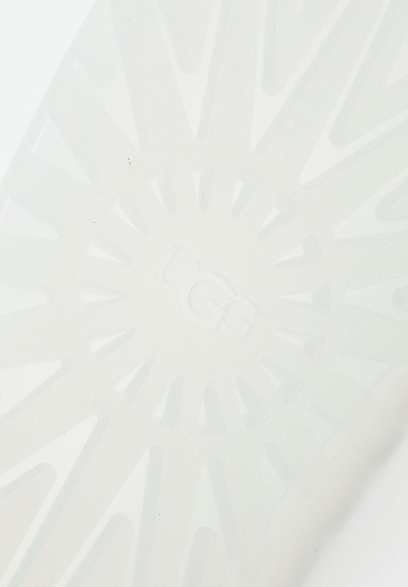 Biała gumowa podeszwa z wzorem w kształcie promieni słonecznych, z wypukłymi rowkami i centralnym, wytłoczonym logo "UGG", zaprojektowana z myślą o trakcjach i trwałości.