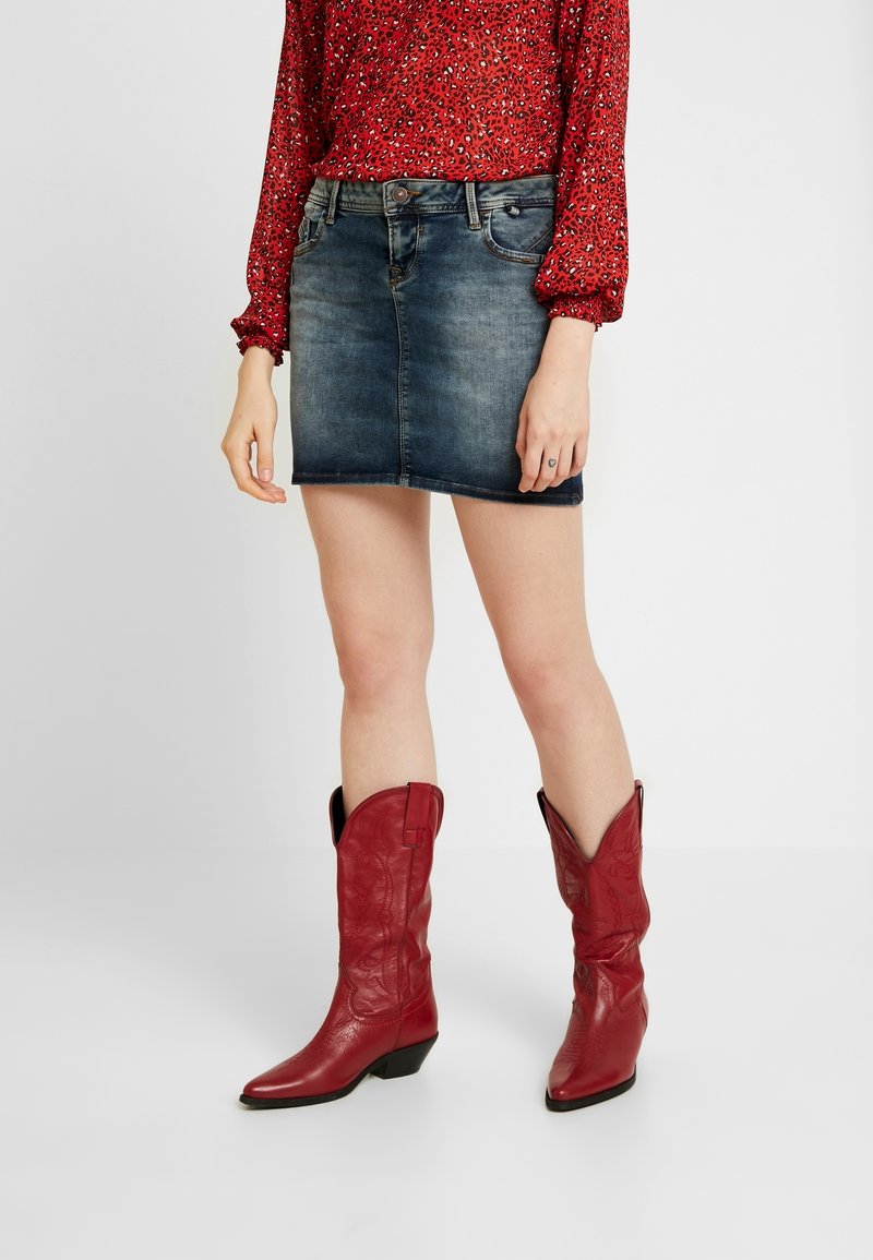 Blusa a maniche lunghe rossa con motivi floreali abbinata a una mini gonna in denim scuro e stivali cowboy rossi in pelle alti fino al ginocchio, con punta affusolata e tacco basso.