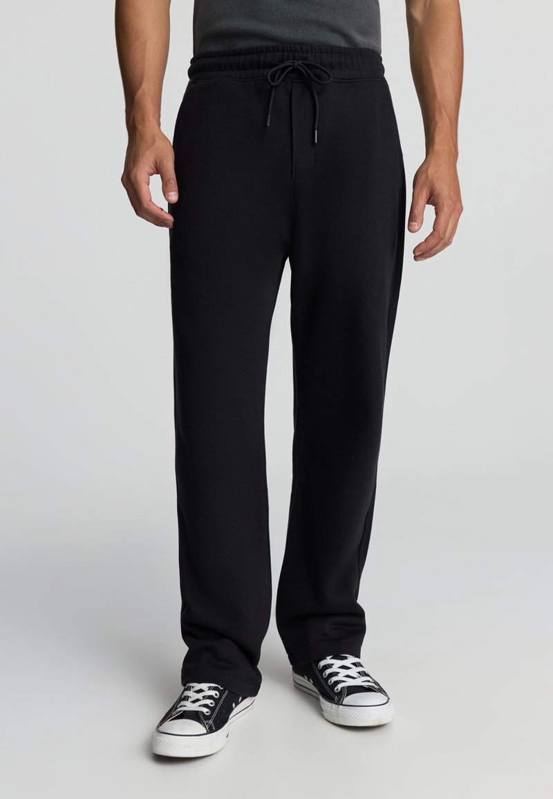 Pantaloni sportivi neri realizzati in tessuto morbido, con vita elastica e cordino, vestibilità rilassata e senza tasche. Abbinati a sneaker nere.