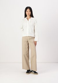 Bela kardigana z vtičenim vzorcem preko srajce s kragnico, v kombinaciji z bež širokimi hlačami in črnimi supergami z belimi črtami.