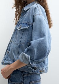 Džínová bunda ve světle modré barvě, s klasickým дизайnem, dvěma náprsními kapsami, zapínáním na knoflíky, ohrnutými rukávy a vybledlou texturou.