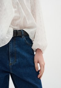 Blusa bianca e testurizzata con righe verticali, abbinata a jeans in denim blu con una cintura nera. Si mette in evidenza il contrasto tra tessuto e colore.