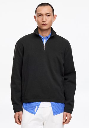 Vyras dėvi juodą ketvirčio užtrauktuko megztinį ant mėlynos dryžuotos marškinės ir baltas kelnes, stovėdamas priešais vientisą baltą foną.