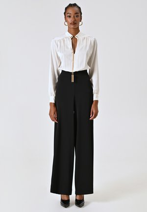 Blusa bianca a maniche lunghe con scollatura profonda e dettagli in oro, abbinata a pantaloni neri a vita alta e ampia gamba con tacchi neri.