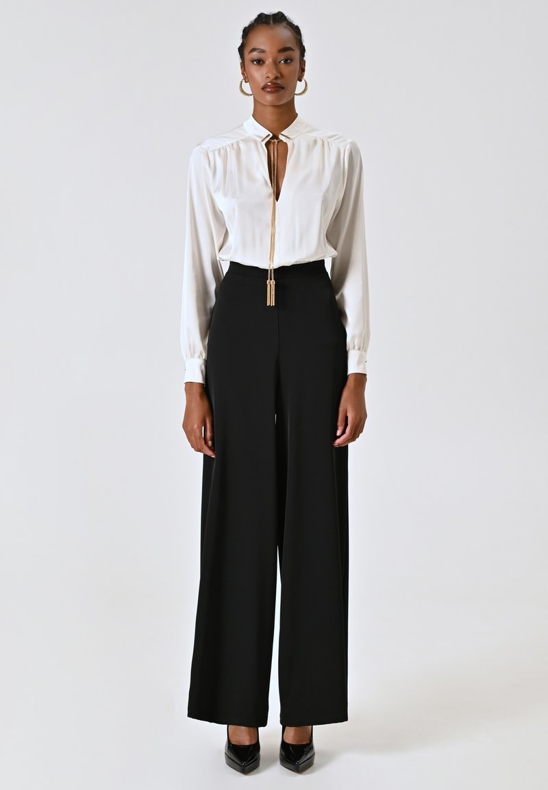 Blusa bianca a maniche lunghe con scollatura profonda e dettagli in oro, abbinata a pantaloni neri a vita alta e ampia gamba con tacchi neri.