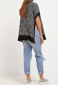 Poncho à motifs noir et blanc avec un ourlet à franges, associé à un jean à taille basse délavé, avec des roulottes et des baskets argentées.