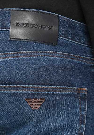 Vaqueros de mezclilla azul oscuro con una etiqueta de cuero en la cintura con el relieve "EMPORIO ARMANI" y un logotipo de águila en tono cobre en el bolsillo trasero.