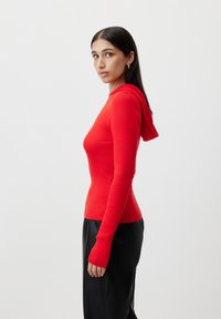 Red ribbed long-sleeve sweater with a fitted shape and a hood. It has thumbholes at the cuffs and is paired with black trousers.